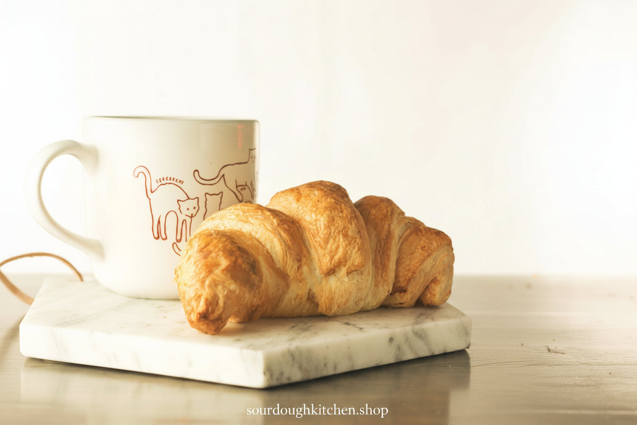 A golden-brown croissant resting on a white marble board next to a white ceramic mug with a minimalist cat line drawing, set on a light wooden table against a bright, warm background.